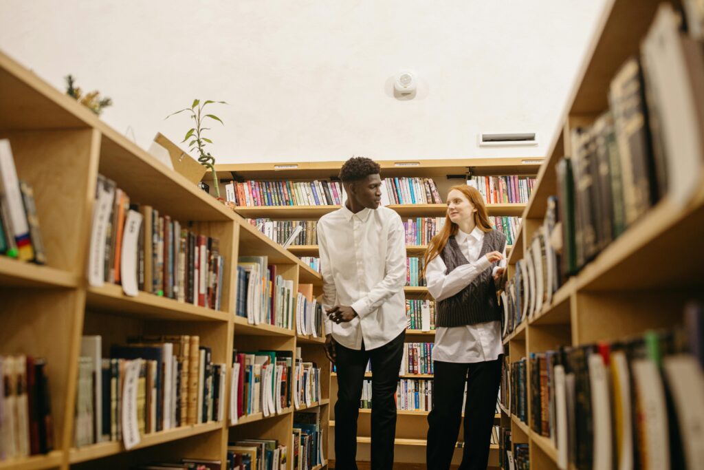 A black man and caucasian woman discussing in a library aisle surrounded by bookshelves.