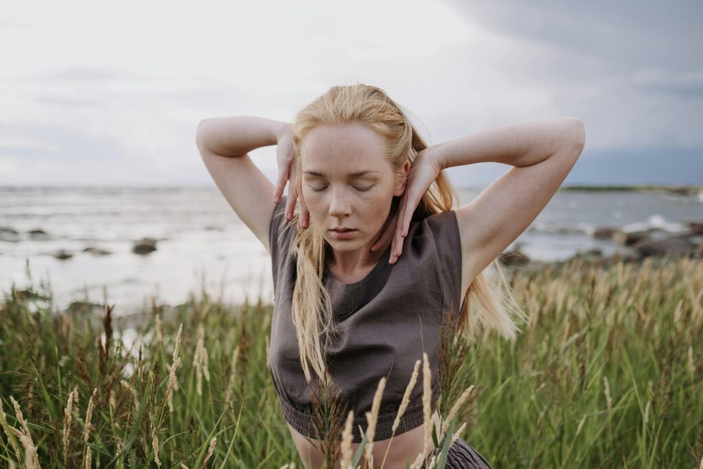 A woman gracefully dances with closed eyes in a coastal grass field, embracing nature.