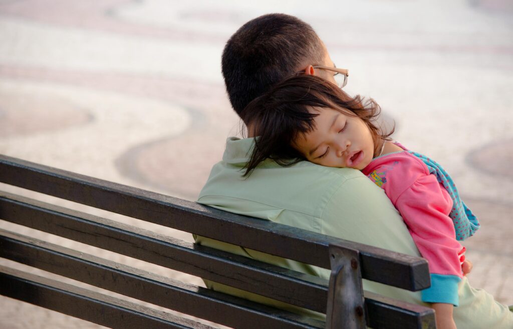 A serene portrait of a sleeping child in a father's arms on a park bench outdoors.
