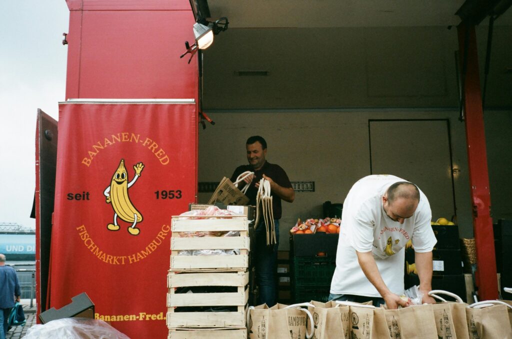 Two workers unloading produce bags at a Hamburg market stand.