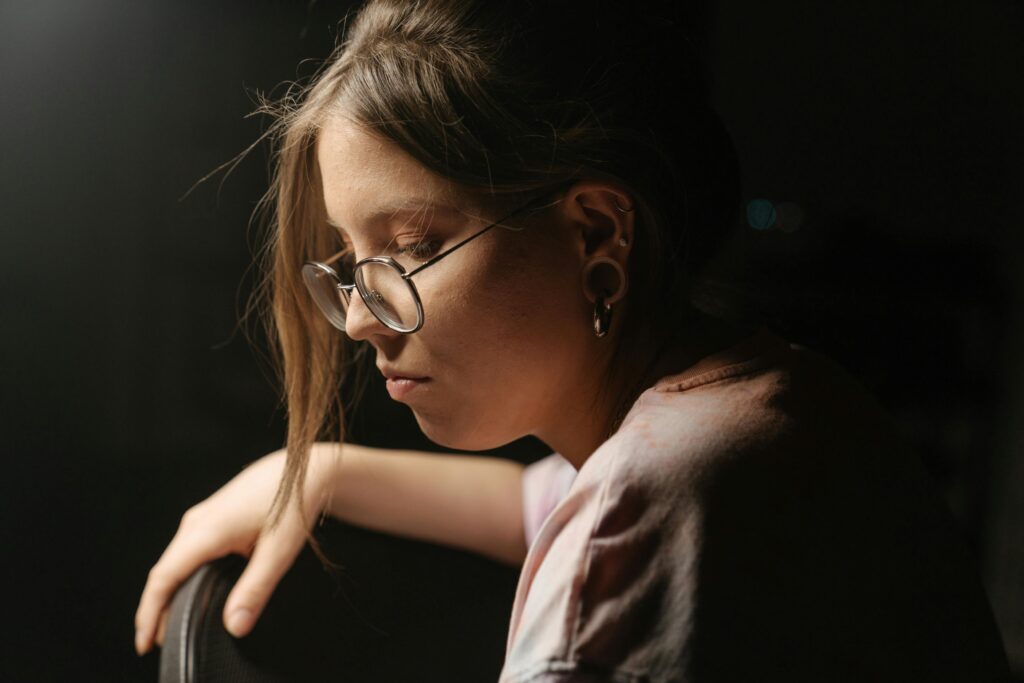 A thoughtful young woman with glasses reflecting quietly in low light indoors.