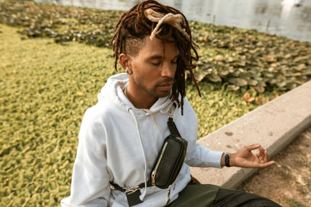 A man with dreadlocks meditates in a park setting, eyes closed in peaceful contemplation.