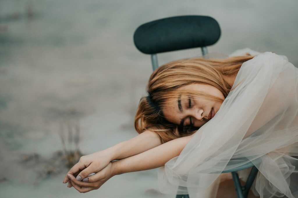 Young dreamy ethnic female with dyed hair and closed eyes covered with transparent fabric on chair