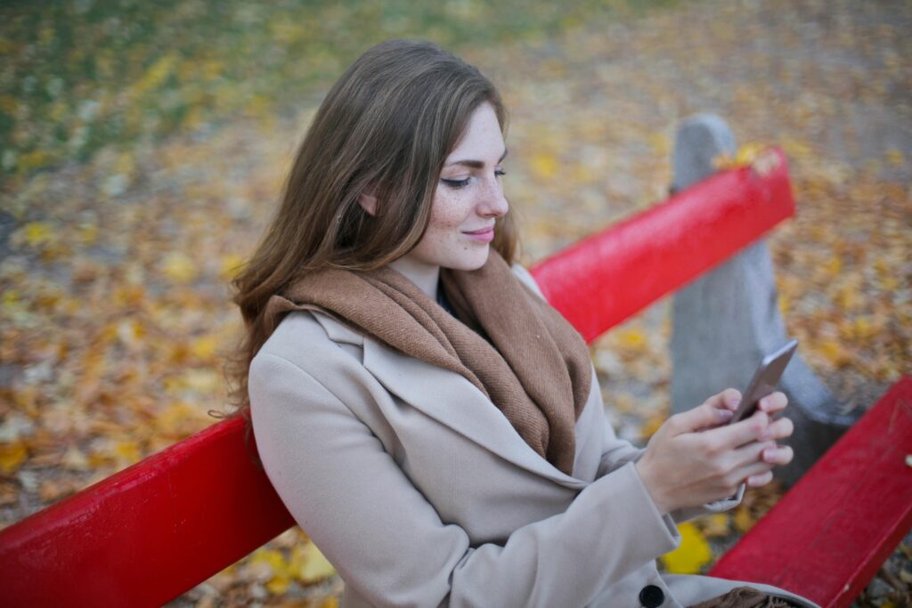 Young woman sitting on a red bench using her smartphone in an autumn park setting.