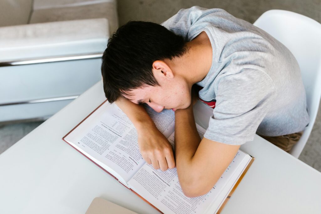 A young man sleeping on a white table with an open textbook, depicting study fatigue.